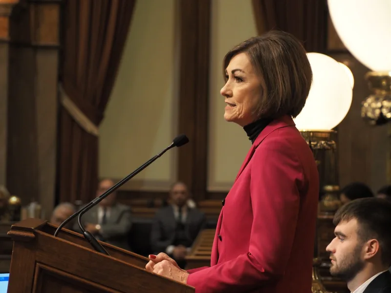 Iowa Gov. Kim Reynolds speaking at an event, wearing a red jacket, with brown curtains and round lights in the background.