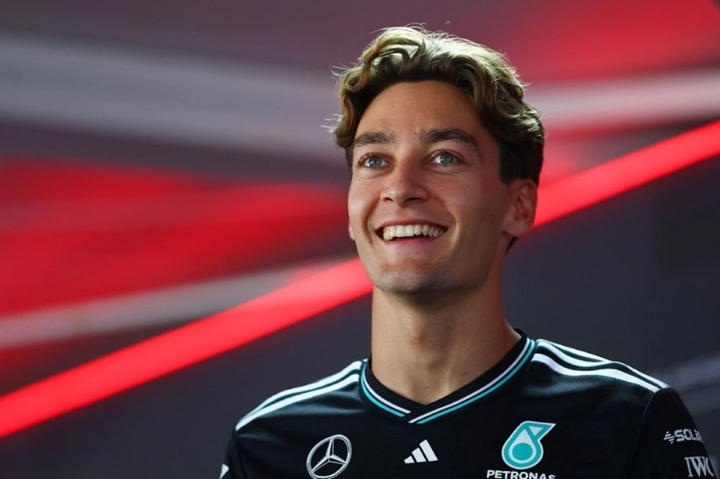 George Russell: smiling in Mercedes F1 team shirt, portrait with red light.