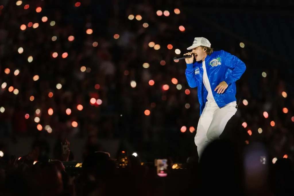 Morgan Wallen sings on stage in blue jacket and cap during 2026 tour, concert crowd lights twinkle, Image Credits: Saskia Potter