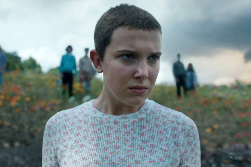 Millie Bobby Brown as Eleven in Stranger Things, standing in field, Image Credits Stranger Things / Netflix