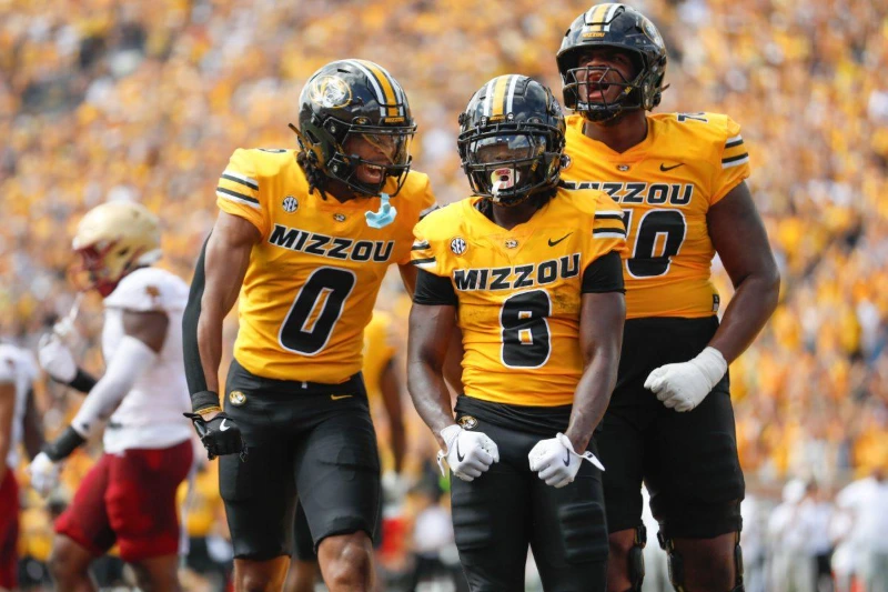 Mizzou football players #0, #8, and #70 united in celebration on field, team spirit.
