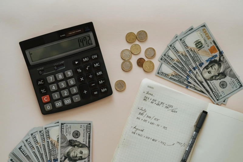 Chartered accountant tools—calculator, US dollar bills, coins, financial notebook with calculations. Image Credits: Olia Danilevich / Pexels