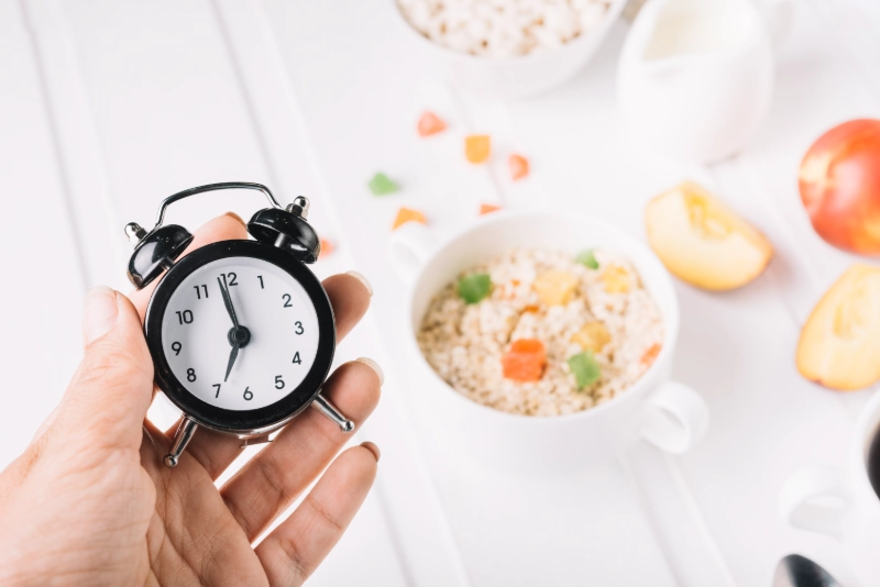 Hand holding clock, healthy breakfast and fruits in background, symbolizing time-restricted eating. Image Credits: FREEPIK