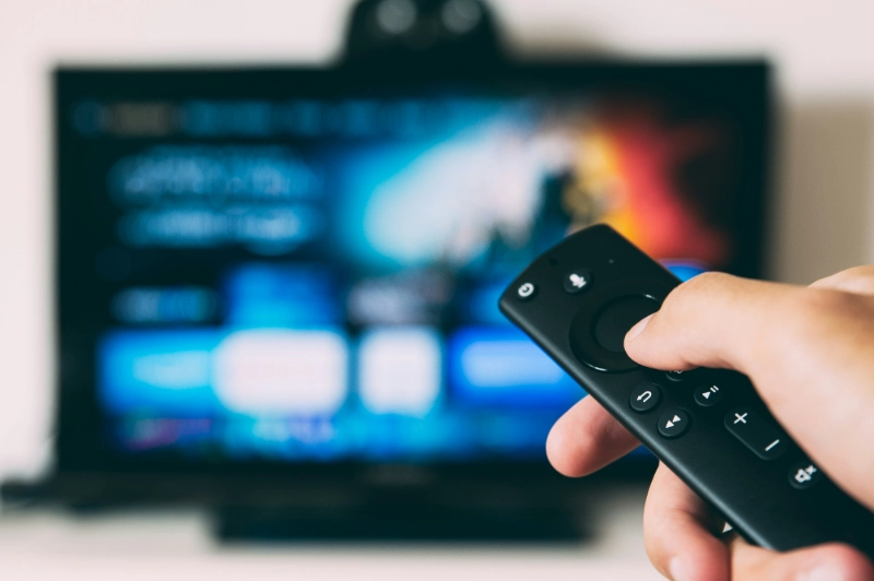 Hand holding TV remote in front of streaming screen, image credits - Glenn Carstens-Peters / UNSPLASH