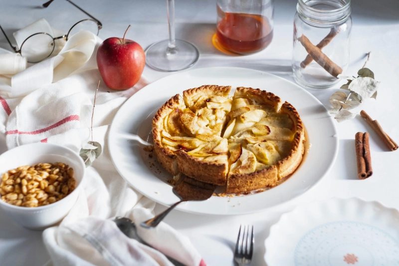 Sliced apple pie on white plate with glass of wine, apple, and cinnamon sticks. Image credits - Diliara Garifullina / UNSPLASH