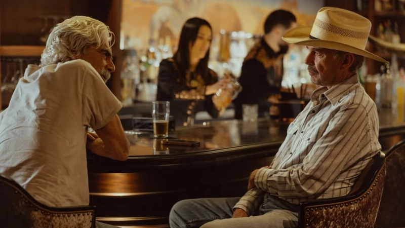 Two men, one in a cowboy hat and striped shirt, sit at a bar conversing with drinks, as bartenders work in the background. Image Credits - Paramount+