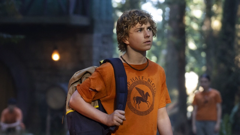 Percy Jackson wears Camp Half-Blood orange shirt and backpack, ready for adventure in the woods. Disney+ series.
