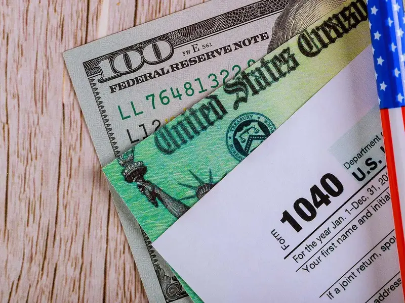 A close-up of a $100 bill, a United States Treasury check, and part of an IRS Form 1040 related to $1400 Stimulus Checks.
