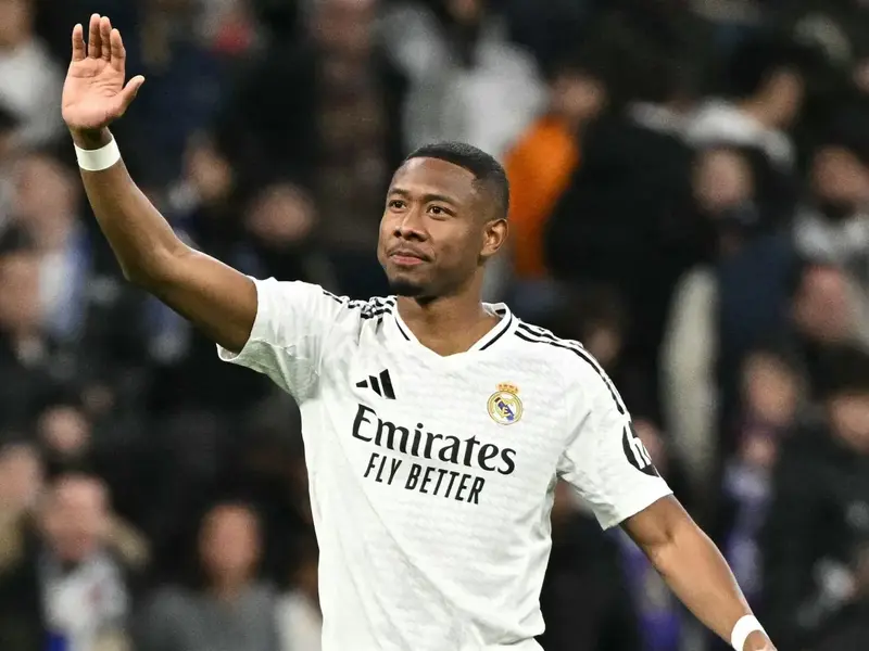 David Alaba soccer player in Real Madrid jersey with black stripes, raising one arm with blurred spectators in the background. Image credit: ©APA/AFP/JAVIER SORIANO."