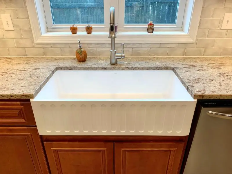 White apron-front deep sink with ribbed detailing, granite countertop, gooseneck faucet, and wooden cabinets beneath a window.