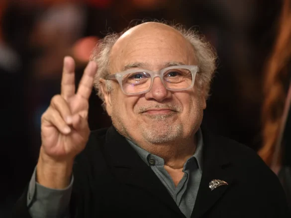 Danny DeVito: peace sign at event, smiling in black jacket.