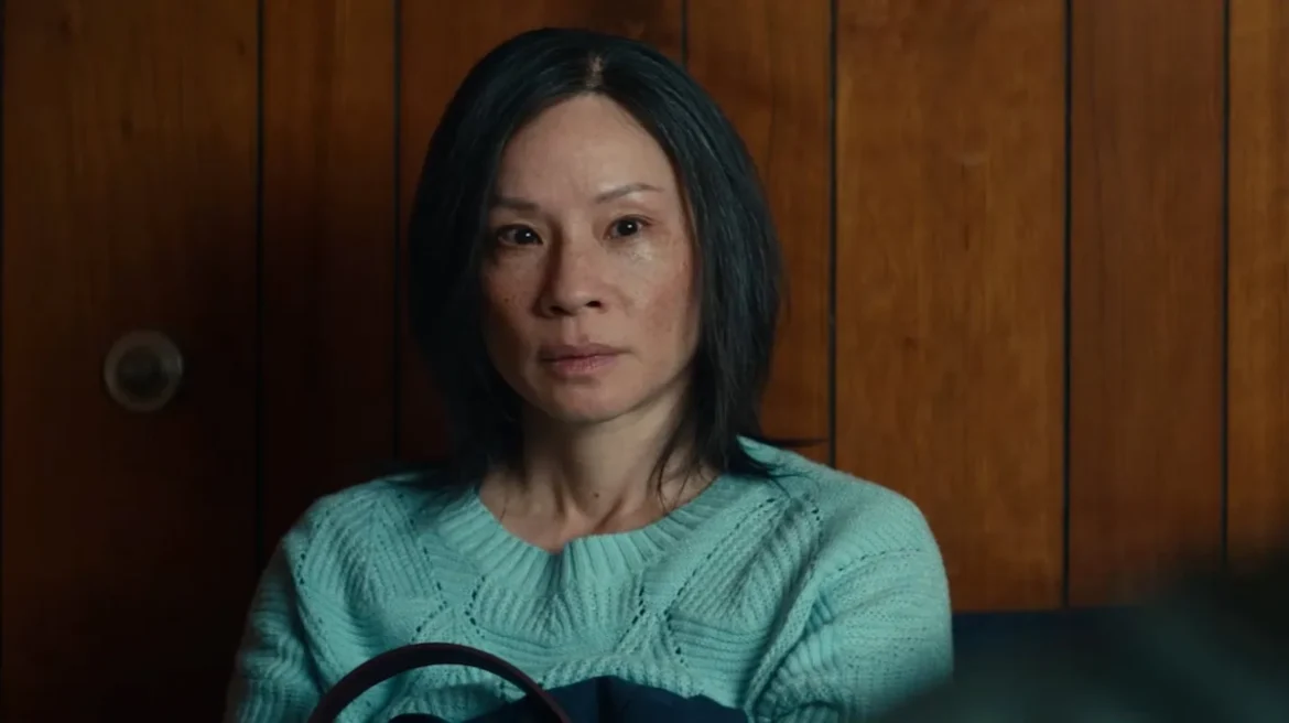 Lucy Liu in a blue sweater, sitting quietly in a wood-paneled room, contemplative mood.